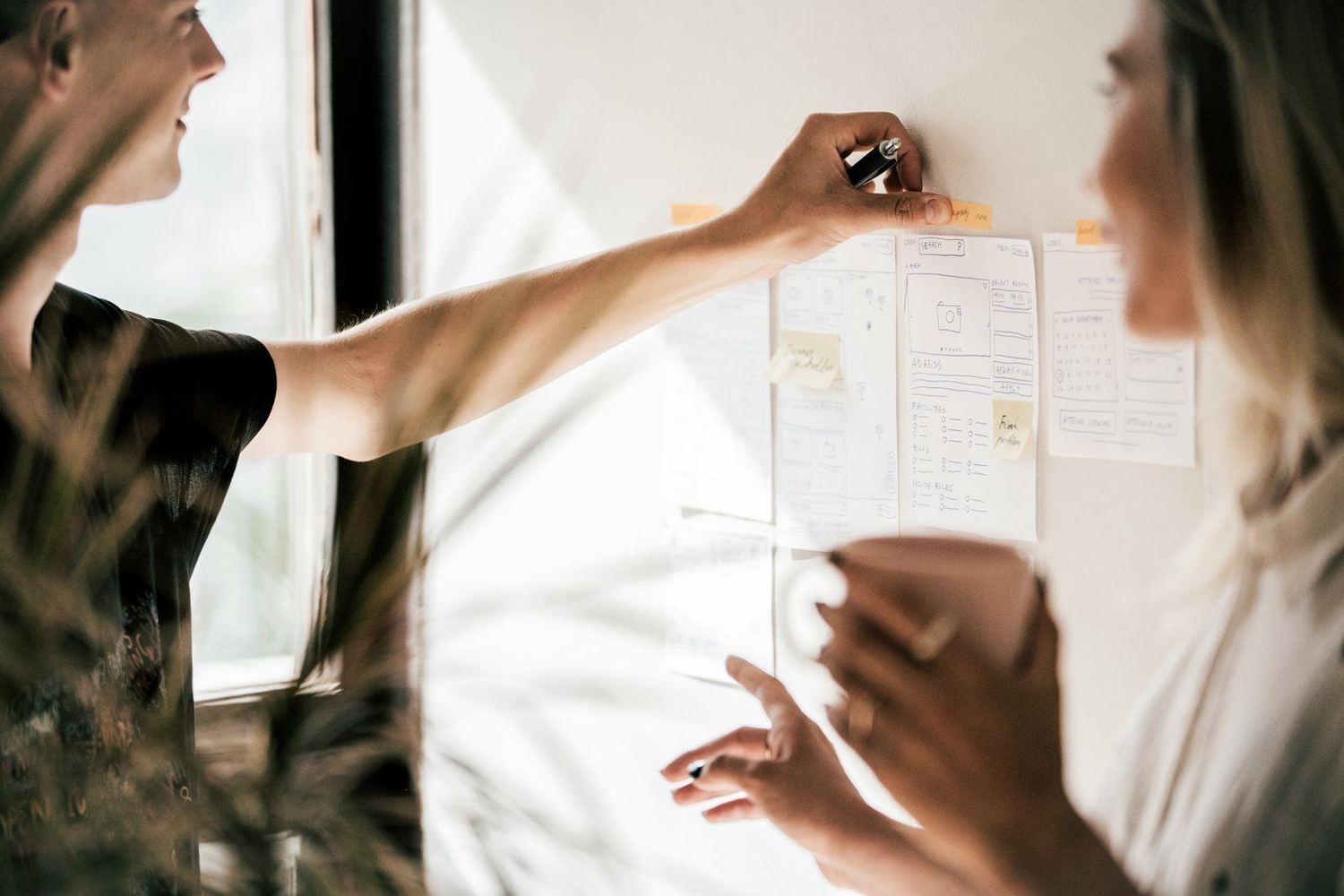 Two people working side by side at a wall of sketches and sticky notes